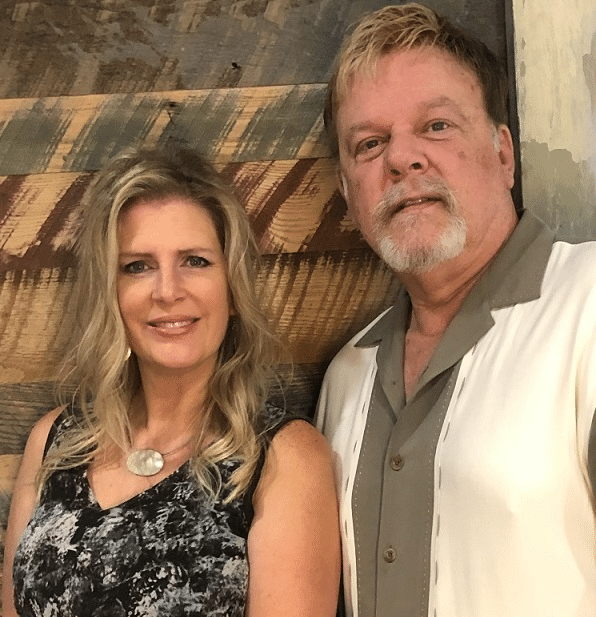 A woman with wavy hair and a stone necklace poses beside a man in a casual shirt, both smiling against a rustic wooden backdrop.